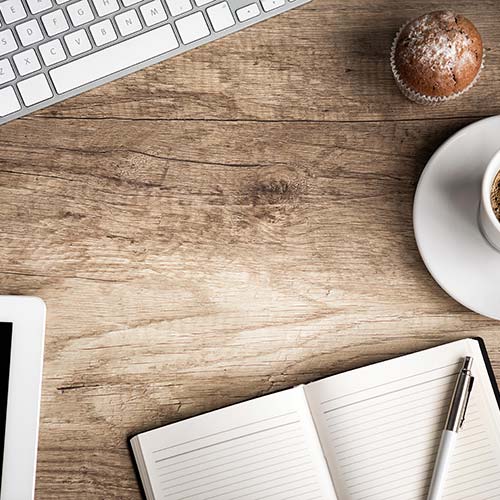 wooden desk with a keyboard, cup of coffee, and notebook on it